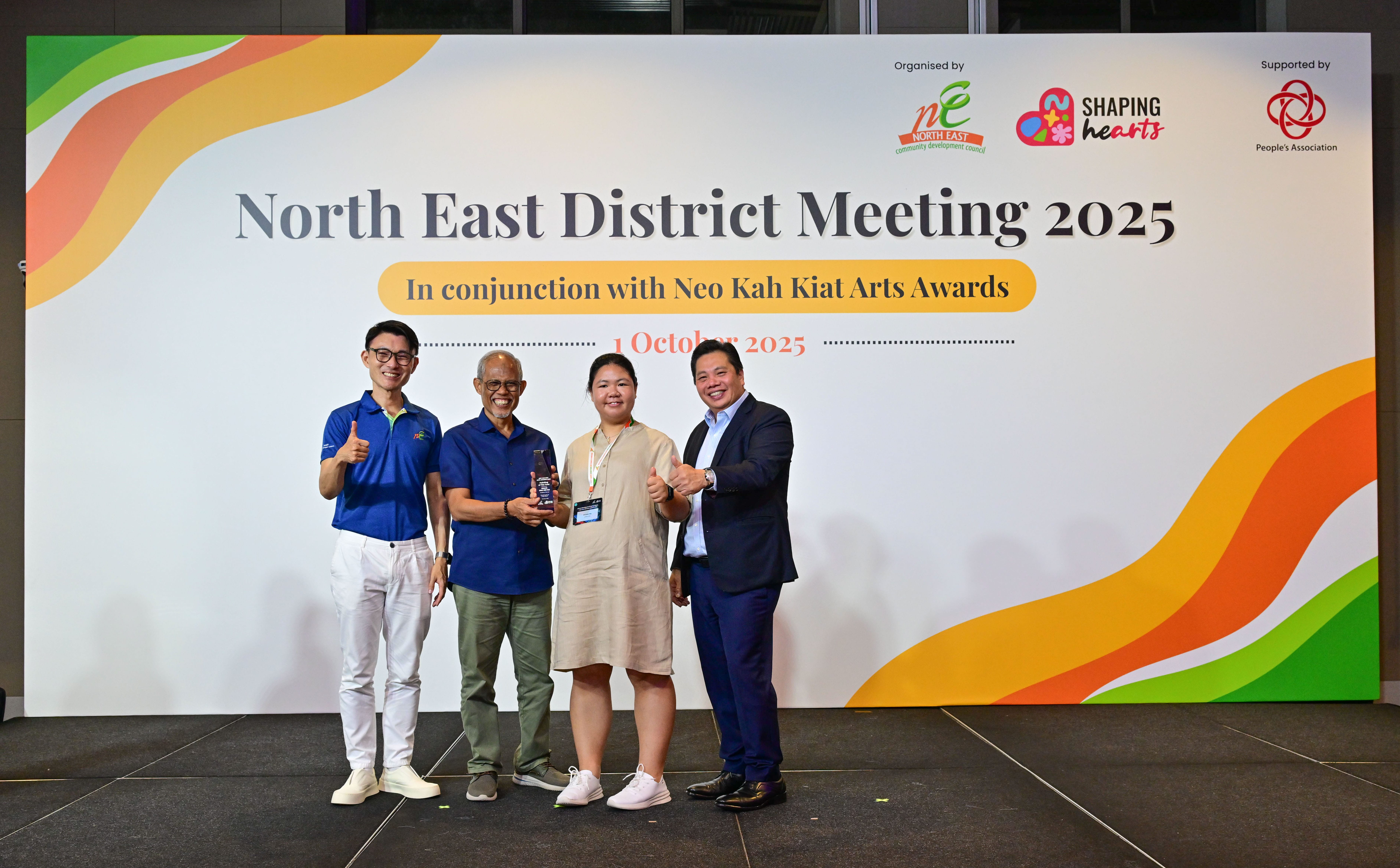Award recipient standing on stage with the presenter, holding a acrylic plaque and posing for a group photo during the award ceremony, with a backdrop reading ‘North East District Meeting 2025 In conjunction with Neo Kah Kiat Arts Awards'